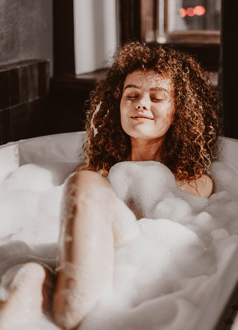 baden_im-winter_schaumbad_hautpflege-rau-cosmetics_b Frau mit lockigem Haar, entspannt in einer Schaumbadewanne, umgeben von schaumigem Wasser, symbolisiert hautfreundliche Winterpflege und Entspannung.