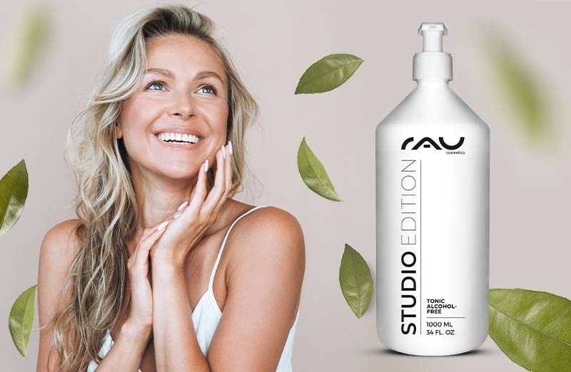 Lächelnde Frau mit RAU Studio Edition Flasche, umgeben von Blättern, präsentiert alkoholfreies Hautpflegeprodukt.